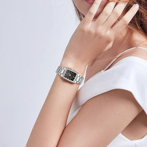 Relógio feminino minimalista pequeno com pulseira prata e visor preto no pulso de uma mulher